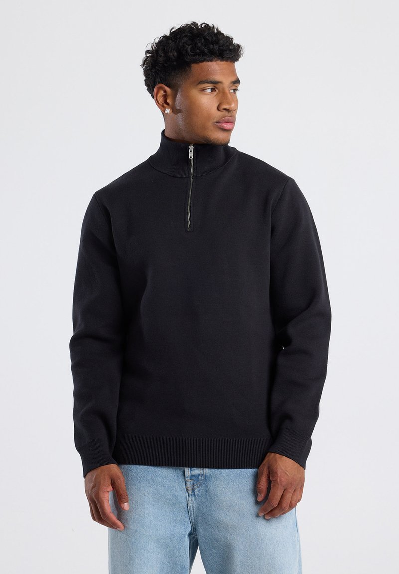 Junger Mann mit kurzen lockigen Haaren, der einen schwarzen Pullover mit halbem Reißverschluss und hellblaue Jeans trägt und seitlich vor einem einfarbigen Hintergrund schaut.