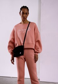 Sweatshirt de algodão rosa claro com o texto "Seja gentil", combinado com calças de fato a condizer. Bolsa de ombro preta com ferragem circular. Textura suave.