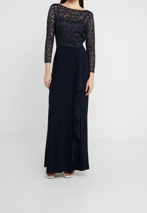 Femme portant une longue robe bleu marine avec des manches longues en dentelle et un détail à volants sur le devant, debout devant un fond clair uni.