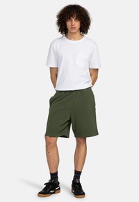 White cotton t-shirt with a chest pocket, paired with green relaxed-fit shorts. Model wears black sneakers and black socks.