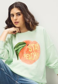 Mäta zelená mikina s ilustráciou broskyne a nápisom "STAY PEACHY" vo žltej farbe. Mäkký materiál a voľný strih, s manžetovými rukávmi.