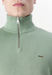 Groene geribbelde half-zip pullover van gestructureerde stof met een zilveren rits. Heeft een klein geborduurd logo op de borst.