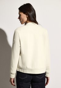 Sweat-shirt de couleur crème avec une texture lisse, des poignets côtelés et un col rond, présentant un écusson logo sur l'épaule gauche.