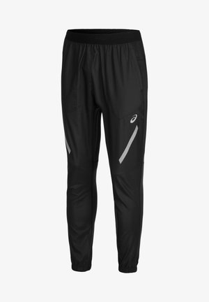 Homme portant un pantalon de sport noir avec des détails réfléchissants et des chaussures de course multicolores, debout devant un fond blanc.