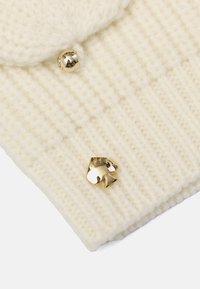 Creme strikket hue med ribbet tekstur, rund guldknapdetalje og et skinnende guld spade-logo foran.