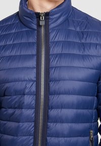 Marineblaue Steppjacke mit hohem Kragen, horizontal gesteppten Details, einem zentralen Reißverschluss und einer seitlichen Reißverschlusstasche.