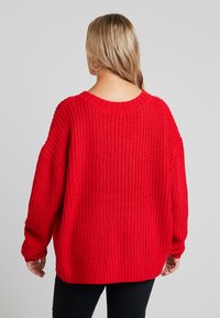 Red oversized knitted sweater with a wide neckline, ribbed cuffs, and textured pattern. The hemline is slightly longer at the back.