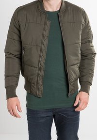 Puffjacka i olivgrön, med dragkedja fram, ribbade ärmslut och quiltad design. Bärs över en mörkgrön T-shirt och jeans.