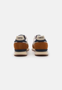Pantofi sport cu un upper bej, toc din suede maro, accente albastre și o talpă căptușită de culoare bej. Numărul "1973" este vizibil pe toc.