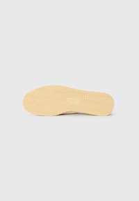 Rubber shoe sole in beige with a zigzag tread pattern. Smooth texture and rounded toe design, featuring a subtle brand logo embossed.