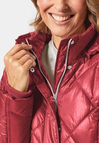 Gequiltede, felrode jas met hoge kraag, zilveren rits en ribgebreide mouwen. Het materiaal lijkt glanzend en lichtgewicht.