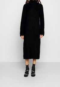 Svart, långärmad stickad klänning med hög krage, med avslappnad passform. Återfinns tillsammans med svarta öppna klackstövlar med metalliska detaljer.