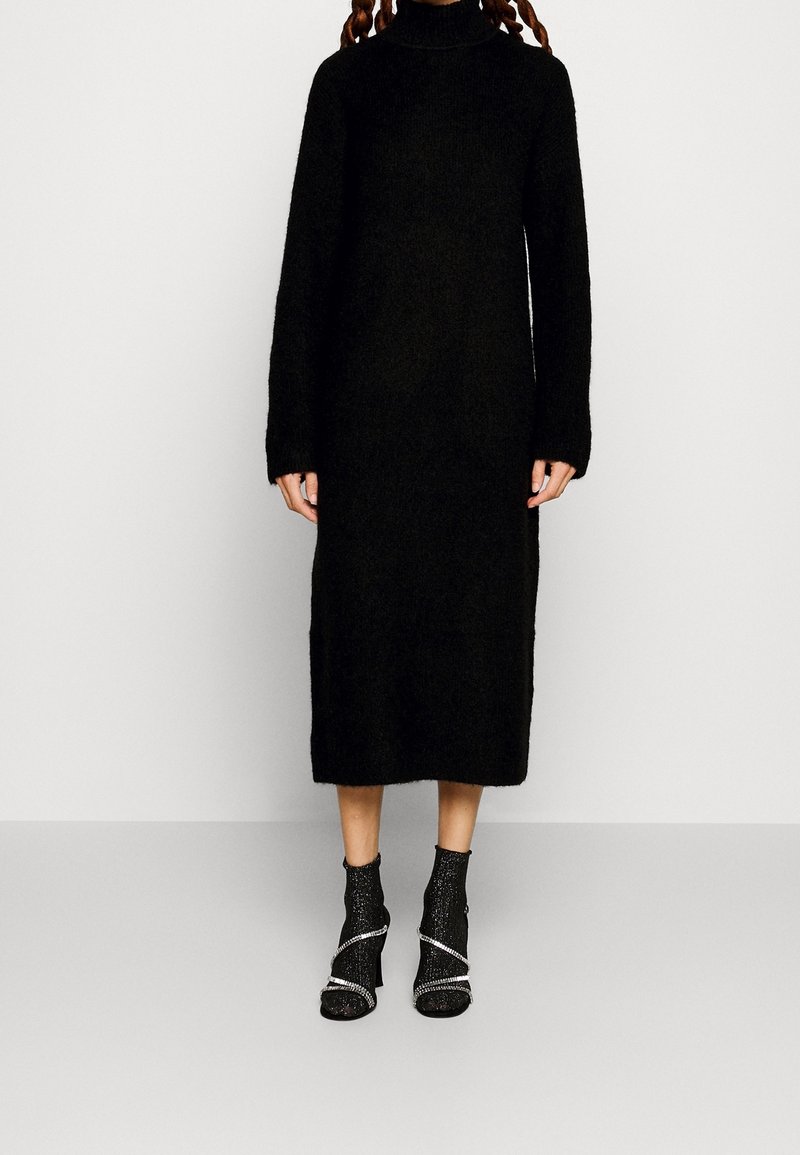 Svart, långärmad stickad klänning med hög krage, med avslappnad passform. Återfinns tillsammans med svarta öppna klackstövlar med metalliska detaljer.
