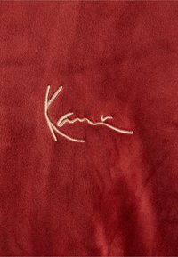 Rød ruskind med en blød tekstur, prydet med et cremefarvet broderet signatur, stiliseret som "Kami" i en flydende skrift.