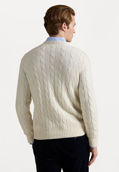 Pull en tricot torsadé ivoire, avec un design texturé et un ourlet côtelé, porté par-dessus une chemise à col bleu clair.