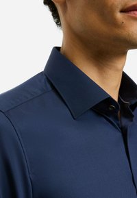 Marineblaue Hemdbluse mit strukturiertem Kragen, glatter Textur und tailliertem Schnitt mit einem einzelnen Knopf nahe dem Ausschnitt.