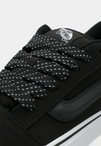 Svart sneaker med en teksturert overdel, flate svart-hvite rutete snører, og et svart lærdetalj nær snorlokkene. Hvit mellomsåle inkludert.