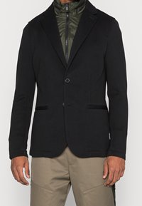 Svart kavaj med en slät yta, enkel knappstängning och två framfickor. Lagd över en olivgrön zippered innerjacka.