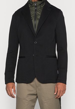 Czarna marynarka o gładkiej fakturze, z pojedynczym zapięciem na guzik oraz dwoma przednimi kieszeniami. Nosi się na oliwkowej zielonej kurtce zapinanej na zamek.