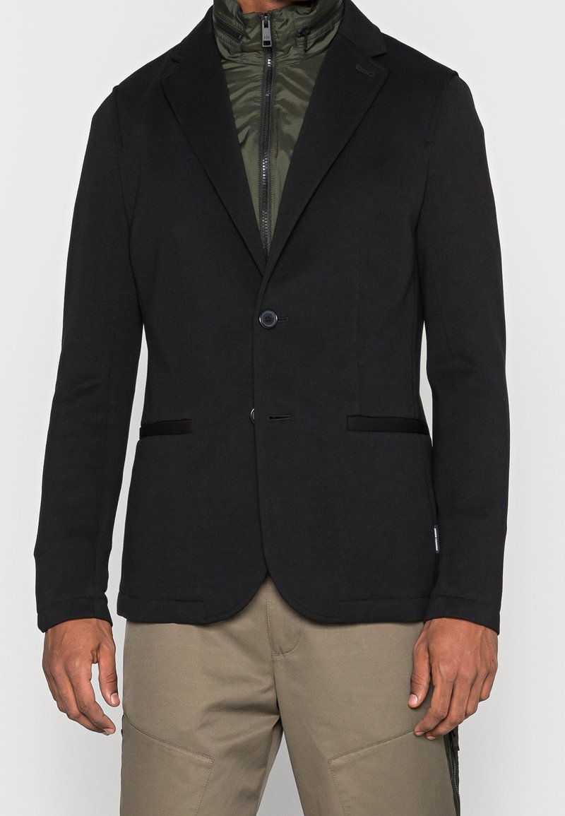 Svart kavaj med en slät yta, enkel knappstängning och två framfickor. Lagd över en olivgrön zippered innerjacka.