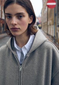 Szary hoodie na zamek z kapturem, wykonany z teksturowanej tkaniny, nałożony na białą koszulę z kołnierzykiem, na tle rozmytej ulicy.