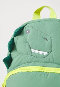 Green dinosaur backpack with quilted texture, featuring embroidered facial details, a yellow zipper, and a bright yellow handle.