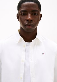 Camisa branca de algodão com botão, com um pequeno logótipo bordado em vermelho, branco e azul. Usada sobre uma t-shirt branca.