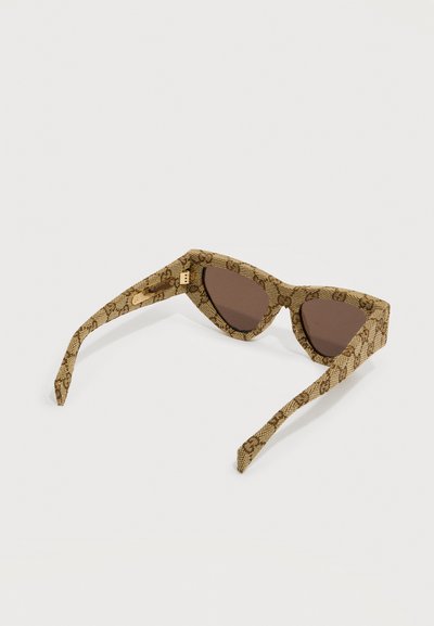 Lunettes de soleil œil de chat avec des verres teintés marron et des montures beiges à motifs posées à plat sur une surface blanche.