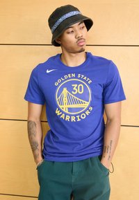 Camiseta azul de Nike con el logo amarillo de los Golden State Warriors y un diseño de puente. El modelo lleva pantalones verdes y un sombrero de cubo negro.