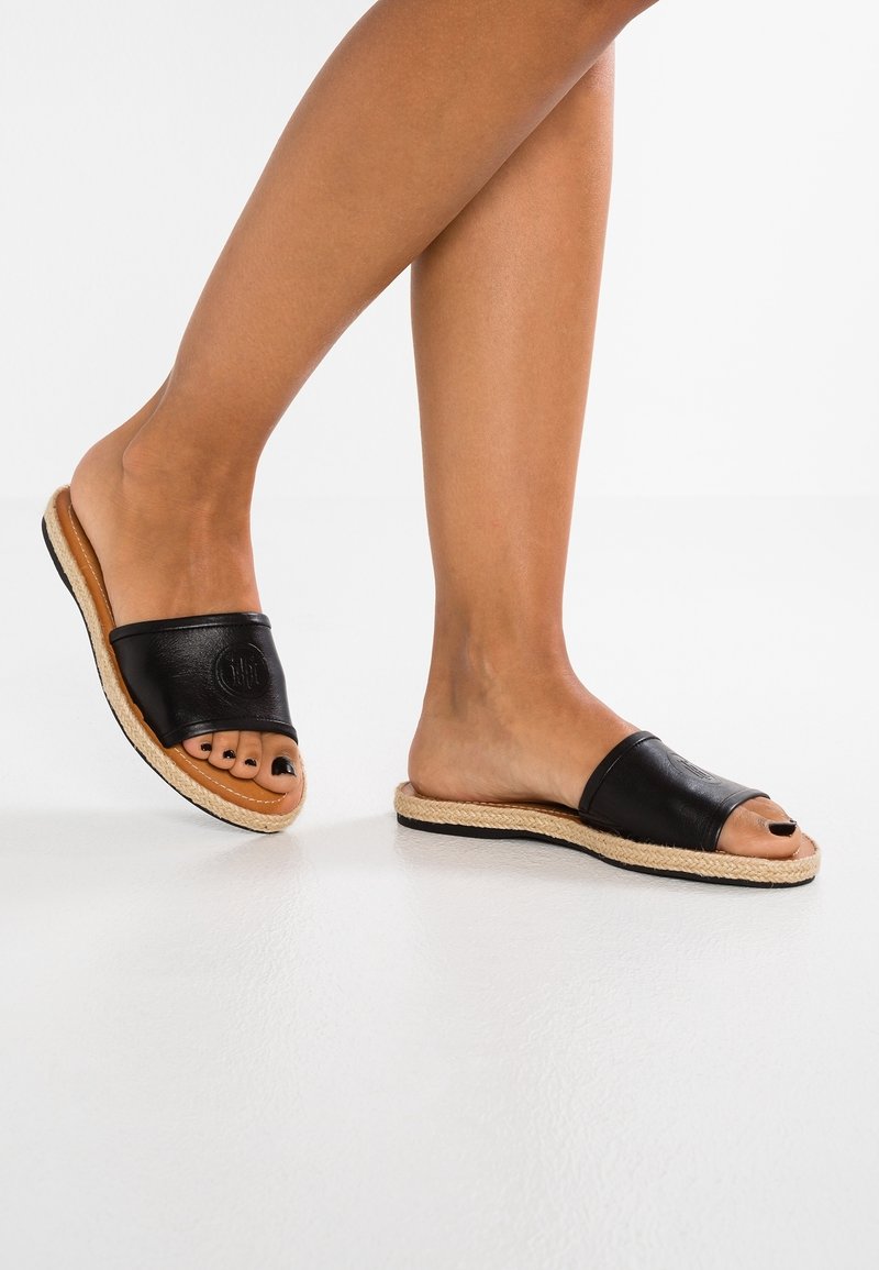 Jambes portant des sandales ouvertes noires à enfiler avec des semelles en corde tressée et des semelles intérieures couleur caramel, debout sur un sol blanc brillant.