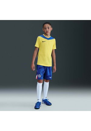 Dreng iført gul og blå fodbolddragt med hvide knæstrømper og blå fodboldstøvler, står mod en mørk gradient baggrund.