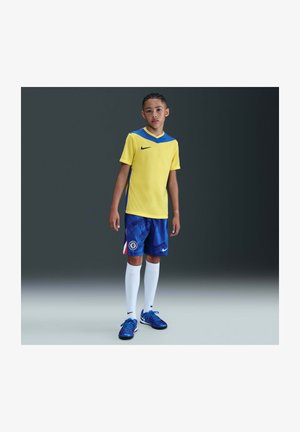 Niño con uniforme de fútbol amarillo y azul, calcetines blancos hasta la rodilla y botas azules, de pie contra un fondo con degradado oscuro.
