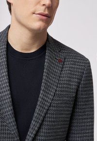 Marineblauer Hahnentrittmantel aus strukturiertem Stoff, verziert mit einem kleinen roten Akzentlogo am Revers, getragen über einem dunklen Strickpullover.