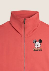 Veste zippée corail avec un col montant. Présente un graphique de Mickey Mouse en sequins sur la poitrine et un tissu lisse et texturé.