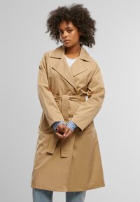 Beige trench coat with a belt, lapel collar, long sleeves, and side pockets, worn over a white shirt with blue striped cuffs.