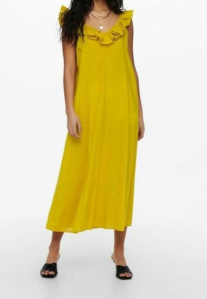Žena v žlutých midi šatech bez rukávů s volánovým výstřihem do V a černých sandálech s otevřenou špičkou stojí před bílým pozadím.