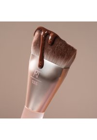Pinceau de maquillage incliné étiqueté RT 135 avec du fond de teint liquide marron dégoulinant de ses poils sur un fond beige.