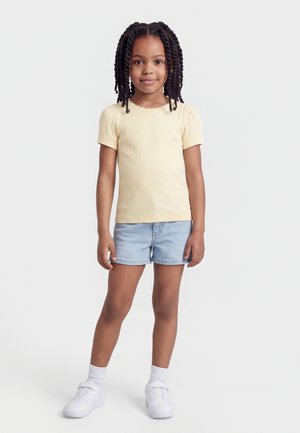 Fille aux cheveux tressés se tenant de face, portant un haut à manches courtes jaune clair, un short en denim clair, des chaussettes blanches et des baskets blanches.