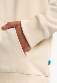 Sudadera beige con un bolsillo frontal, puños acanalados y una pequeña etiqueta turquesa en el lateral, hecha de un suave tejido texturizado.