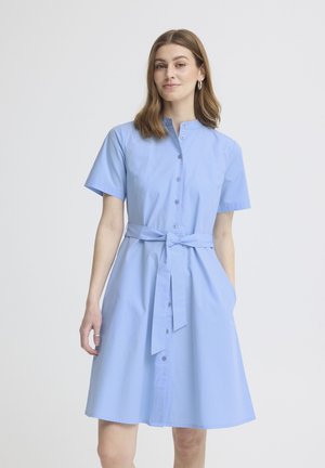 Mladá žena v svetlomodrých šatách so zapínaním na gombíky, krátkymi rukávmi a opaskom s viazaním v páse, stojaca pred jednoduchým bielym pozadím.