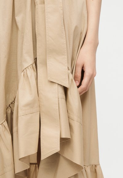 Χέρι που ακουμπά δίπλα σε μπεζ ασύμμετρη φούστα με βολάν και φαρδιά ζώνη σε απλό φόντο.