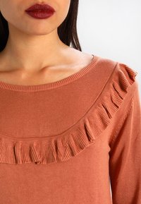 Haut couleur rouille avec une texture côtelée, présentant un large décolleté et des volants décoratifs le long du bord. Tissu lisse, coupe ajustée.