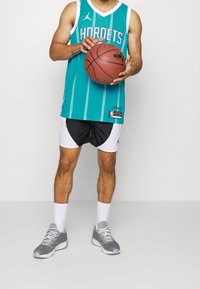 Teal koripallo paita valkoisilla raidoilla ja "HORNETS" -teksti, yhdistettynä mustiin ja valkoisiin shortseihin, harmaisiin lenkkikenkiin, pitelee koripalloa.