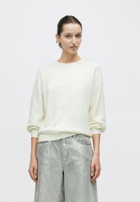 Ανοιχτό κρεμ φούτερ με στρογγυλή λαιμόκοψη και μανίκια τύπου νυχτερίδας, με ανάγλυφο λογότυπο στο κέντρο. Συνδυασμένο με πλατύσελα μεταλλικά ασημί παντελόνια.