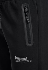 Pantalons de sport noirs avec une texture lisse, dotés d'une poche zippée avec un accent en caoutchouc et d'un logo imprimé en blanc sur le côté.