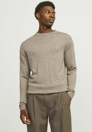 Hellbrauner Pullover aus weichem Strickmaterial, mit Rundhalsausschnitt, langen Ärmeln und schmaler Passform. Kombiniert mit beigen Hosen, die Falten und Taschen haben.