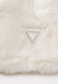 Matériau en fausse fourrure de couleur blanc cassé avec une texture douce. Présente une étiquette triangulaire en argent frappée du mot "GUESS" et d'un point d'interrogation.
