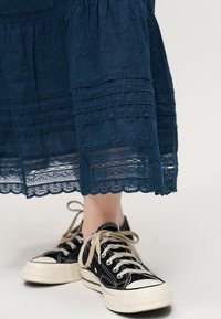 Jambe inférieure portant des baskets à lacets noires et blanches associées à une jupe bleu marine ornée de détails brodés et d'un ourlet en dentelle.