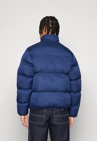 Ναυτικό μπλε μπουφάν με οριζόντια σχέδια, υψηλό γιακά και λεία, γυαλιστερή επιφάνεια. Φοριέται πάνω από σκούρο τζιν.