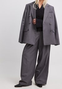 Grauer doppelt geknöpfter Blazer aus strukturiertem Stoff, dazu passende weit geschnittene Hose und ein schwarzes Hemd darunter. Schwarze Schuhe runden das Outfit ab.